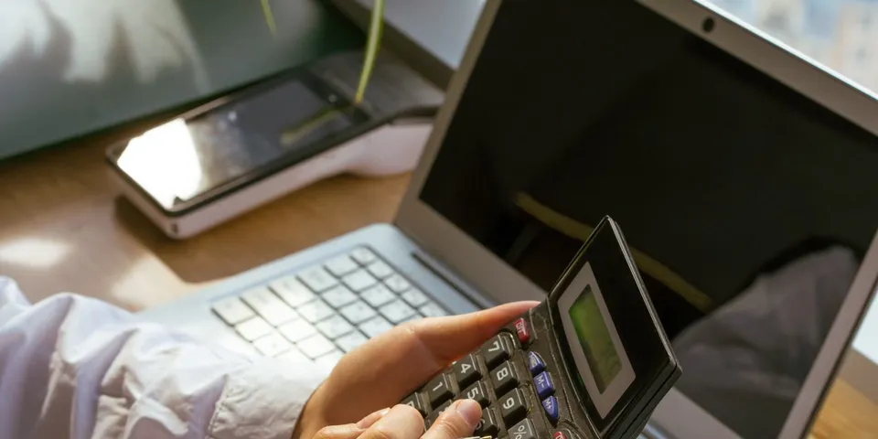 Person kalkuliert an einem Büroarbeitsplatz mit Laptop und Taschenrechner die laufenden IT-Kosten eines Unternehmens